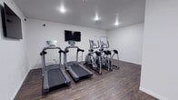 This single-floor fitness room offers a straightforward and functional workout space designed for cardiovascular exercise. The room features two treadmills and two elliptical machines, each equipped with digital displays to help users track their workout metrics. A wall-mounted flat-screen television is positioned directly in front of the cardio equipment, allowing residents or guests to stay entertained during exercise sessions.

The space is finished with durable dark hardwood-style flooring, which provides both an attractive aesthetic and a practical surface suited to fitness equipment. The walls are painted a clean, bright white, contributing to an open and airy atmosphere despite the room's modest footprint. Recessed ceiling lights illuminate the space evenly, ensuring a well-lit environment during any time of day.

The minimalist design keeps the layout uncluttered, maximizing usable floor space around the equipment. A whiteboard is mounted on one wall, which may serve as a space for posting schedules or fitness information. Overall, the room presents a clean, no-frills environment well-suited for residents seeking convenient access to essential cardio fitness equipment.
