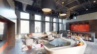 This sophisticated single-floor lounge space showcases a striking contemporary design that blends industrial and modern aesthetics. Soaring ceilings with exposed ductwork and dark finishes are complemented by an eye-catching lighting installation featuring oversized cylindrical pendant lamps suspended within delicate brass halo rings, creating a dramatic overhead focal point. Floor-to-ceiling windows line the perimeter, flooding the interior with abundant natural light while framing views of the surrounding urban cityscape. The main lounge area is thoughtfully arranged with plush, neutral-toned sofas and curved accent chairs, inviting conversation and relaxed gatherings. Vibrant geometric-patterned area rugs in warm terracotta, ivory, and charcoal tones anchor the seating clusters and inject a bold design element underfoot. A sleek bar or reception counter finished in richly veined marble stone anchors the rear of the space, backed by a bold red feature wall that serves as a vivid accent. Wood-and-metal bar stools add a casual elegance to this zone. Throughout the space, a carefully curated mix of low-profile coffee tables in muted tones balances the energy of the colorful textiles, resulting in an environment that feels both vibrant and refined — ideal as a communal gathering or hospitality venue.