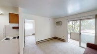 Located at 122 Stoddard Ave in East Lansing, Michigan, this single-floor unit offers a functional and straightforward layout spread across several connected living spaces. The living room is carpeted and finished in neutral white tones, featuring baseboard heating, a wall-mounted air conditioning unit, and a sliding glass door that opens to a private balcony overlooking mature trees, bringing in natural light and greenery. The adjoining dining area transitions to hardwood-style flooring and includes a ceiling fan overhead, a closet with bi-fold doors, and a seamless connection to the kitchen. The kitchen is equipped with warm-toned wood cabinetry and a sink, providing a compact and practical cooking space. The bedroom is carpeted and well-lit by a window with vertical blinds that frame views of surrounding trees, complemented by baseboard heating for year-round comfort. The bathroom features white tile work, a vanity with dark cabinetry, and a shower-tub combination. Throughout the unit, the clean white walls and neutral palette create a simple, cohesive atmosphere suited for everyday living.