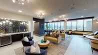 This elegantly designed high-rise amenity lounge occupies a single open floor and showcases a seamless blend of contemporary style and refined comfort. The space is anchored by expansive floor-to-ceiling windows that wrap around the perimeter, flooding the interior with natural light and offering sweeping panoramic views of the surrounding cityscape.

The lounge is organized into several distinct yet connected zones. A central seating area features plush velvet sofas and accent chairs in deep navy and gold tones, arranged around a marble-topped round coffee table and layered over a patterned area rug. Decorative multi-globe chandeliers illuminate the space with warm, ambient light throughout.

A dedicated wet bar area, finished with glossy black subway tile backsplash and a beverage refrigerator, provides a functional and stylish entertaining station. Nearby, a brick accent wall adds textural contrast and grounds one end of the lounge, complemented by a wall-mounted flat-screen television.

A separate dining nook features a stunning live-edge wood conference table surrounded by upholstered quilted chairs, bathed in natural light from corner windows with city views on two sides — ideal for private gatherings or meetings. Additional high-top bar seating along the windows offers casual vantage points overlooking the skyline.

A bold navy geometric accent wall with integrated lighting adds depth and a distinctive design focal point to the relaxation corner, complete with leather chaise lounges. Light porcelain tile flooring ties the entire space together with a clean, modern finish.