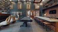 Located at 1121 3rd Ave N in Nashville, TN, this single-floor amenity lounge delivers an impressive blend of industrial character and modern sophistication. Soaring ceilings define the space, amplifying the sense of openness across each distinct zone.

The media lounge area anchors one end of the space, featuring a massive wall-mounted display framed in warm wood cabinetry, complemented by a sectional sofa, eclectic natural wood coffee tables, and a patterned area rug in earthy tones. Floor-to-ceiling windows flood the area with natural light and offer views of the surrounding courtyard and pool.

At the heart of the space sits a full-size billiards table with a matte black finish, surrounded by woven lounge chairs and bench seating. Exposed brick walls, wood-framed transom windows, and globe pendant lighting lend the area a distinctly Nashville-inspired, warehouse aesthetic. A decorative slatted wood screen partition adds architectural interest.

The wet bar area is outfitted with a substantial concrete-top island, built-in cabinetry in rich wood tones, dual wall-mounted displays, and eclectic industrial wall art, creating an ideal space for social gatherings.

A comfortable seating nook with a neutral sofa, copper-toned accents, and framed French doors leading to adjacent spaces completes this well-appointed communal environment.