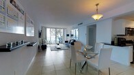 This single-floor residence features an open-concept layout that seamlessly integrates the living and dining areas into one cohesive, well-lit space. The interior is finished in a clean, contemporary style, with light-toned large-format tile flooring running throughout, contributing to a sense of spaciousness and continuity.

The dining area is anchored by a circular glass-top table surrounded by sleek white chairs, illuminated by a warm pendant light overhead. Adjacent to it, the living area is furnished with a navy blue sofa and complementary accent pieces arranged atop a charcoal area rug, adding texture and contrast against the predominantly white palette.

Floor-to-ceiling windows along the far wall flood the interior with natural light and frame views of the outdoors, including what appears to be a balcony and greenery beyond. The white walls and ceilings enhance the airy, open feel of the space. Additional details include recessed ventilation, a wall-mounted television, and neutral drapery framing the expansive glazing. The overall ambiance is clean, modern, and bright.