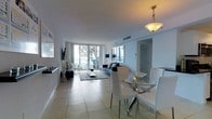 This single-floor residence features an open-concept layout that seamlessly integrates the living and dining areas into one cohesive, well-lit space. The interior is finished in a clean, contemporary style, with light-toned large-format tile flooring running throughout, contributing to a sense of spaciousness and continuity.

The dining area is anchored by a circular glass-top table surrounded by sleek white chairs, illuminated by a warm pendant light overhead. Adjacent to it, the living area is furnished with a navy blue sofa and complementary accent pieces arranged atop a charcoal area rug, adding texture and contrast against the predominantly white palette.

Floor-to-ceiling windows along the far wall flood the interior with natural light and frame views of the outdoors, including what appears to be a balcony and greenery beyond. The white walls and ceilings enhance the airy, open feel of the space. Additional details include recessed ventilation, a wall-mounted television, and neutral drapery framing the expansive glazing. The overall ambiance is clean, modern, and bright.