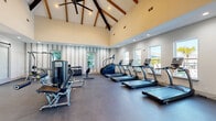 Located at 1201 Cleveland Ave in Wildwood, Florida, this fitness center offers a comprehensive workout facility spread across a single level. The space is divided into two distinct areas designed to accommodate a wide range of exercise needs.

The main workout hall is an architectural standout, featuring dramatically vaulted ceilings with exposed wooden beams and ceiling fans that add both character and airflow to the space. Large windows line the walls, flooding the room with natural light and offering views of the surrounding outdoor areas, including glimpses of a pool. The durable dark concrete flooring supports a robust lineup of equipment, including multiple treadmills, ellipticals, stair climbers, cable machines, free weights, and adjustable benches.

A secondary studio room provides a more intimate setting with light hardwood flooring, a wall-mounted flat-screen TV set into a textured tile accent wall, a large mirror, and a ceiling fan. This area is ideal for spin cycling and focused training sessions. A well-stocked accessory wall features medicine balls, resistance bands, and balance equipment.

Connecting the two spaces is a transitional corridor with a stylish sliding barn door, adding a modern design touch. Natural light permeates throughout, creating a bright and motivating environment well-suited for a full fitness experience.
