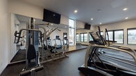 Located at 930 W 17th Ave in Junction City, Oregon, this single-level fitness center is designed to support a comprehensive workout experience. The space features a clean, modern aesthetic with neutral white walls, recessed lighting, and durable rubber flooring throughout, providing both functionality and a professional gym atmosphere.

The facility is organized to accommodate a variety of training styles. The cardio zone includes multiple treadmills and an elliptical trainer, positioned near large windows that allow natural light to fill the space and offer views of the surrounding landscape. Wall-mounted flat-screen televisions are positioned throughout for entertainment during workouts.

The strength training area is equipped with a Hoist multi-station cable machine, providing a range of resistance exercise options. A dedicated free weights section includes a full dumbbell rack with a weight bench, situated beside a bank of windows that brings in additional natural light. Floor-to-ceiling mirrors along one wall visually expand the space and assist with form during exercise.

Additional amenities include a wall-mounted water fountain, a wall-mounted mini-split HVAC unit for climate control, and a glass-paneled door providing access to the space. The overall layout is well-organized, offering an efficient and comfortable environment for fitness activities.
