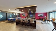 This dynamic single-floor amenity space is thoughtfully designed to support a variety of social and recreational activities. The centerpiece of the space is a fully equipped communal kitchen, anchored by a striking dark marble island with built-in gas cooktop, stainless steel oven, and a bold wood-paneled ceiling detail with integrated range hood. Bold magenta accent walls and vibrant artwork lend the kitchen and dining area an energetic, eclectic character, complemented by upholstered bar seating along the island. Adjacent dining tables with mixed modern chairs offer flexible seating arrangements. Large sliding glass doors open directly to an outdoor patio, flooding the interior with natural light and providing seamless indoor-outdoor connectivity. The game and lounge area features a moody teal palette, a shuffleboard table, plush sectional and accent seating, and a neon sign creating an inviting, social atmosphere. A dedicated screening room with a large-format display, built-in shelving, and deep navy walls offers a more intimate retreat for viewing. The space also includes an accessible restroom and light wood-look tile flooring throughout, unifying the various zones with a clean, contemporary foundation.