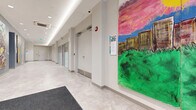 This single-floor commercial space presents a clean, contemporary design characterized by light-toned walls, neutral tile flooring, and recessed lighting that creates a bright, well-illuminated environment throughout. The layout comprises interconnected corridors and functional zones that together form an organized, accessible facility.

The main entrance opens into a welcoming lobby featuring glass entry doors that invite natural light, a seating area with a light blue sofa and round coffee tables, and curated artwork displayed on the walls. Poetic text inscribed directly on the wall adds a thoughtful, community-oriented touch to the entry experience.

Extending from the lobby, wide corridors connect the various areas of the space. These hallways are distinguished by large-scale murals and paintings — vibrant, expressive works depicting landscapes, abstract forms, and community themes — transforming circulation paths into a gallery-like experience.

An SSM Health Express Clinic operates within the space, indicated by prominent signage near the corridor. Additional rooms with frosted glass partitions line the hallways, providing private or semi-private functional areas. Safety features including fire extinguisher cabinets and alarm systems are integrated throughout.

A dedicated bike storage room offers multi-tier bicycle racks, accommodating numerous bikes efficiently. The overall ambiance balances clinical functionality with an arts-forward, community-conscious character.