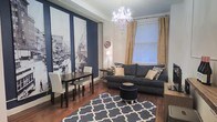 This thoughtfully designed single-floor space makes a striking first impression with its bold navy blue accent wall, which serves as a dramatic backdrop for large-scale black-and-white cityscape artwork. The combination of deep tones and classic imagery lends the space a sophisticated, urban character.

The living area is anchored by rich hardwood flooring and a navy geometric-patterned area rug, creating a cohesive color palette throughout. A plush charcoal sofa, accented with gold and patterned throw pillows, offers comfortable seating and is complemented by a sleek round ottoman that doubles as a functional centerpiece. Natural light filters in through a large window dressed in elegant gold curtains, brightening the space and enhancing its warm ambiance.

Overhead, a crystal chandelier adds a touch of glamour and serves as a standout architectural feature, elevating the overall aesthetic of the room. A compact dining area with a dark wood table and white chairs is neatly integrated into the space, making efficient use of the layout while maintaining a polished look.

The neutral white ceiling and walls provide a clean contrast to the bold design choices, ensuring the space feels balanced and inviting. Overall, this space successfully blends classic elegance with contemporary style, creating an atmosphere that is both visually engaging and functionally comfortable.