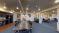 This well-appointed single-floor communal space is thoughtfully designed to accommodate a variety of gathering and work-related activities. The layout is open and expansive, anchored by a substantial rustic wood conference table surrounded by dark spindle-back chairs — an ideal centerpiece for meetings, group dining, or collaborative sessions. Recessed lighting throughout provides consistent, warm illumination across the carpeted floors.

A comfortable lounge area features modern upholstered seating, including a striped accent sofa and sleek side chairs, creating an inviting spot for casual conversation or relaxation. Natural light filters in through horizontal-blind-covered windows along one wall, adding brightness to the neutral-toned interior.

A smaller breakout area with a compact table and task chairs offers a quieter setting for focused work or small group discussions, accentuated by framed artwork on the walls.

The kitchenette is fully equipped with dark cabinetry, an electric range, microwave, dishwasher, refrigerator, and a sink with extended counter space — all finished with warm hardwood flooring that contrasts nicely with the surrounding carpet. A private office or workroom is accessible through a separate door, fitted with a desk and task chairs for individual use. The overall ambiance is clean, functional, and welcoming.