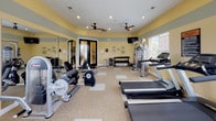 This fitness center is designed to support a well-rounded workout routine, offering a comprehensive selection of equipment within a single, open room. The space is finished with warm yellow and soft gray accent walls, complemented by recessed lighting and dual ceiling fans that maintain air circulation throughout. A patterned carpet-and-tile flooring provides both comfort and durability underfoot.

Large windows fitted with white shutters line multiple walls, allowing generous natural light to filter in while offering views of the surrounding outdoor greenery. A wall-mounted flat-screen television is positioned for easy viewing during workouts.

The cardio section includes multiple treadmills and an elliptical machine, arranged to take advantage of the natural light from the windows. A cable crossover machine and additional strength equipment provide options for resistance training, while a rack of dumbbells in varying weights caters to free weight exercises. Medicine balls on a dedicated storage rack offer further versatility.

Full-length mirrors along one wall visually expand the space and allow users to monitor their form. The fitness policies are prominently posted, and the overall layout ensures ample room to move between stations comfortably.