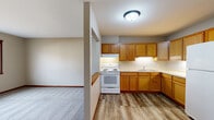 Located at 2003 9th St E in Menomonie, Wisconsin, this one-bedroom, one-bathroom unit offers a practical and well-organized single-floor layout. The kitchen is a standout feature, designed in an efficient L-shape with warm honey-toned oak cabinetry extending from floor to ceiling, light countertops, a double-basin sink, and white appliances including a range and refrigerator. Durable wood-look flooring runs throughout the kitchen, lending a clean and cohesive look. A half-wall partition with a wood ledge separates the kitchen from the living area, which transitions to plush carpeting and features a window that draws in natural light and provides views of the surrounding neighborhood. The bedroom is a comfortable, carpeted space with neutral walls, a large sliding-door closet offering ample storage, a window for natural light, and baseboard heating. The bathroom includes a wood-vanity with a single sink, light countertop, a medicine cabinet with mirror, and coordinating wood-look flooring consistent with the rest of the unit. A connecting hallway links the rooms efficiently. Throughout the unit, warm wood tones in the trim, doors, and cabinetry complement the neutral gray walls, creating a functional and inviting living environment.