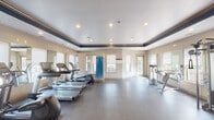 This fitness center offers a spacious, single-floor layout designed to accommodate a full range of workout needs. The facility features a distinctive architectural detail: a tray ceiling accented with a bold navy blue border and warm wood crown molding, lending the space a polished, upscale character. Recessed lighting throughout ensures the room is bright and well-illuminated at all times.

The cardio section is generously equipped with multiple treadmills, elliptical machines, and a stair climber, all arranged to take advantage of natural light streaming through several large windows fitted with white roller shades. A wall-mounted flat-screen television is also present for entertainment during workouts.

The strength training area includes a multi-station cable machine, various selectorized machines, a dumbbell rack with a full set of free weights, and an adjustable workout bench. Large mirrors line multiple walls throughout the space, visually expanding the room and allowing users to monitor their form during exercise. A yoga mat storage unit is conveniently positioned within the space.

Durable, dark rubber flooring covers the entire facility, providing both safety and comfort underfoot. French-style glass doors at the entrance add an elegant touch and allow views into the space. The overall ambiance is clean, modern, and functional, reflecting a commitment to providing a comprehensive and comfortable fitness environment.