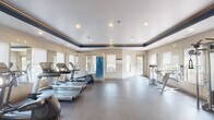 This fitness center offers a spacious, single-floor layout designed to accommodate a full range of workout needs. The facility features a distinctive architectural detail: a tray ceiling accented with a bold navy blue border and warm wood crown molding, lending the space a polished, upscale character. Recessed lighting throughout ensures the room is bright and well-illuminated at all times.

The cardio section is generously equipped with multiple treadmills, elliptical machines, and a stair climber, all arranged to take advantage of natural light streaming through several large windows fitted with white roller shades. A wall-mounted flat-screen television is also present for entertainment during workouts.

The strength training area includes a multi-station cable machine, various selectorized machines, a dumbbell rack with a full set of free weights, and an adjustable workout bench. Large mirrors line multiple walls throughout the space, visually expanding the room and allowing users to monitor their form during exercise. A yoga mat storage unit is conveniently positioned within the space.

Durable, dark rubber flooring covers the entire facility, providing both safety and comfort underfoot. French-style glass doors at the entrance add an elegant touch and allow views into the space. The overall ambiance is clean, modern, and functional, reflecting a commitment to providing a comprehensive and comfortable fitness environment.