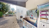 This compact outdoor pet washing station is a purpose-built amenity designed to make pet care straightforward and efficient. The station is sheltered by a covered overhead structure that provides protection from the elements while keeping the area open and airy. The ground is finished with gravel, offering a practical, low-maintenance surface well-suited to an outdoor washing environment.

The centerpiece of the space is a raised stainless steel dog wash tub, which is both durable and easy to clean. The tub is adorned with a playful cartoon graphic of a dog being bathed, adding a lighthearted, welcoming character to the station. A flexible hose attachment is connected to the unit, providing a convenient water source for washing pets. Nearby, a wall-mounted spray or rinse unit offers additional functionality, and a water bowl is placed on the gravel ground for pet use.

Surrounding the station are textured stucco walls and utility enclosures, maintaining a tidy and organized appearance. The open-air design allows natural light to filter in, creating a bright and functional workspace. The station is thoughtfully equipped with posted instructional signage to guide users through proper use of the facilities, making it an accessible and user-friendly amenity for pet owners.