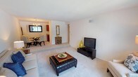 This single-floor residence offers a thoughtfully designed layout that balances comfort and functionality. The living room serves as the heart of the home, featuring neutral-toned carpeting, clean white walls, and an open feel that flows seamlessly into the dining area. The dining space is distinguished by an elegant gold chandelier overhead and a glass-top table, creating an intimate setting for meals. Adjacent to the dining area, the galley-style kitchen is equipped with rich cherry wood cabinetry, stone-look countertops, white appliances including a gas range, and a striking black-and-white checkered tile floor that adds a classic touch. A convenient pantry or utility closet is accessible directly from the kitchen. The bedroom is a serene retreat, offering plush carpeting, bright natural light from a large window with vertical blinds, and a generous reach-in closet with built-in rod and shelving. The full bathroom features tile surround, a tub-shower combination with a patterned curtain, a vanity with sink, and warm pendant lighting complemented by a large mirror. Throughout the residence, a consistent neutral palette and soft carpeting create a cohesive and welcoming ambiance.