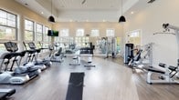Located at 105 Exchange Dr. in Asheville, NC, this single-floor fitness center offers a comprehensive workout environment designed to accommodate a wide range of exercise preferences. The main workout area is expansive and airy, featuring soaring ceilings with a white shiplap finish, pendant lighting, and large windows that flood the space with natural light while providing views of the surrounding outdoor amenity areas, including a pool. Durable hardwood-style flooring runs throughout, and full-length mirrors along the walls enhance the sense of openness.

The main floor is outfitted with a robust selection of TRUE-brand cardio equipment, including treadmills, ellipticals, stationary bikes, and recumbent bikes, all positioned along the windowed walls to maximize views. A dedicated free-weight section includes a full rack of dumbbells paired with flat benches and adjustable training stations, while multiple cable and strength machines provide targeted resistance training options. Wall-mounted televisions are positioned throughout for entertainment during workouts.

A separate spin studio houses multiple red cycling bikes alongside wall-mounted TVs and mirrors, creating a focused environment for cycling classes. The entry area features additional strength equipment and glass doors that connect to outdoor bike storage, reinforcing the facility's active, wellness-oriented character.
