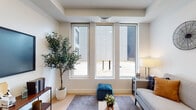 Located at 34 Munroe in Lynn, Massachusetts, this thoughtfully designed single-floor residence offers a clean, modern aesthetic throughout. The living area is bathed in natural light courtesy of a bank of tall, floor-to-ceiling windows that frame outdoor views and lend an open, airy feel to the space. Light hardwood flooring flows continuously throughout, tying each room together cohesively.

The kitchen is well-appointed with gray shaker cabinetry, stainless steel appliances including a refrigerator, range, and microwave, a textured tile backsplash, and a spacious white waterfall-edge island with an undermount sink and pendant lighting overhead — ideal for both cooking and casual dining.

The bedroom provides a calm, restful retreat with warm, neutral tones and ample space. A secondary room functions as a flexible den or home office, featuring its own window for natural light and room for a dedicated workspace with shelving. A full bathroom includes a soaking tub with tiled surround, a floating vanity with an LED-backlit mirror, and the added convenience of an in-unit stacked washer and dryer. Clean white walls, minimalist finishes, and a consistent modern design language define the overall ambiance of this well-planned Lynn residence.