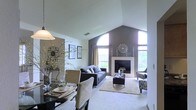 This single-floor residence offers an inviting open-concept layout that seamlessly connects the dining and living areas, creating a cohesive and functional living environment. The living room is the architectural centerpiece of the home, boasting soaring vaulted ceilings that draw the eye upward toward a striking arched window flanked by additional tall windows. This arrangement floods the space with abundant natural light and lends a grand sense of scale to the room. A classic fireplace anchors the far wall, accented by a neutral surround and decorative mantel details, making it a natural focal point for relaxation and gathering. The accent wall in a rich charcoal tone adds depth and contrast against the crisp white walls and ceiling throughout. Plush wall-to-wall carpeting in the living area contributes to a warm and comfortable ambiance. The dining area is positioned adjacent to the living space, defined by elegant pendant lighting and a refined dining set. Framed wall art and decorative botanical accents complete the aesthetic, lending a polished yet welcoming character to the interior. Overall, the home presents a well-balanced blend of architectural interest and thoughtful design throughout its single level.