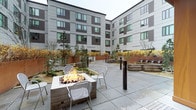 This inviting outdoor courtyard is nestled within a contemporary multi-story residential building, offering residents a well-designed communal gathering space. The centerpiece of the courtyard is a square fire pit surrounded by sleek metal chairs, creating a warm and welcoming focal point for socializing or relaxing. The broad stone-paved patio provides ample room for seating arrangements and easy movement throughout the space.

Lush raised planters line the perimeter, filled with ornamental trees and shrubs that add seasonal color and a natural contrast to the surrounding modern architecture. Slim bollard lighting is thoughtfully positioned throughout the courtyard, ensuring the space is functional and atmospheric after dark.

The enclosing building features a striking façade of dark brown and light gray paneling, with large windows arranged in a regular grid pattern that frame views of the courtyard from multiple floors above. The overall design reflects a clean, modern aesthetic with an emphasis on community-oriented outdoor living.

The courtyard's layout balances open hardscape with softscape elements, offering a sheltered yet open-air environment. Whether used for casual conversation, quiet reflection, or community events, this well-appointed outdoor space serves as a welcoming extension of the residential experience.