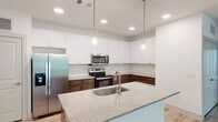 This well-designed single-floor residence offers a clean, contemporary aesthetic throughout. The open-concept kitchen serves as a central highlight, showcasing a two-tone cabinet design with rich walnut lower cabinets and crisp white upper cabinets, complemented by a geometric tile backsplash. A large granite-topped island with an undermount sink and pendant lighting anchors the space, while stainless steel appliances — including a French door refrigerator, electric range, and over-the-range microwave — complete the functional layout. Light wood-look flooring flows through the main living areas, contributing to a bright and airy atmosphere. The living area is generously proportioned, featuring a ceiling fan, natural light from multiple windows, and direct access to an outdoor area via a glass door. The bedrooms are finished with plush neutral carpeting and ceiling fans, providing comfort and warmth. The bathrooms are tastefully appointed with granite vanity countertops and dark wood cabinetry. One bathroom features a glass-enclosed walk-in shower with tile surround, while another offers an elongated vanity with ample storage. Track and recessed lighting throughout the space ensures consistent, well-distributed illumination. The overall design is cohesive, modern, and move-in ready.