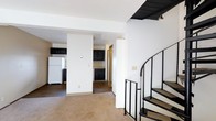 This two-story residence offers a thoughtfully laid-out living environment across two connected floors. The main level features an open living area finished with plush carpeting, creating a warm and comfortable atmosphere. Adjacent to the living space, the kitchen is outfitted with dark, rich cabinetry paired with a tile backsplash, providing ample storage and a classic contrast against the white walls and appliances, including a full-size refrigerator. The kitchen flows seamlessly from the main living area, making the space feel cohesive and functional. A defining architectural feature of the home is the elegant spiral staircase with a sleek black metal railing, which serves as both a practical connection to the upper floor and a visual centerpiece of the interior. The staircase rises gracefully from the main level, drawing the eye upward and adding a distinctive design element to the space. White walls throughout the home contribute to a bright, clean aesthetic, while dark hardwood-style flooring in the kitchen and dining areas complements the cabinetry. The layout balances open communal areas with more defined functional zones, offering a practical and visually appealing living environment across both floors.