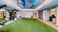Located at 22 Via Toscana in Salem, NH, this professionally designed fitness center spans a generous single floor, offering a comprehensive workout environment divided into two distinct zones. The main cardio and strength area features a modern aesthetic with patterned tile-style flooring in soft blue and grey tones, sleek linear LED lighting, and a central media wall with a large flat-screen TV framed by built-in cabinetry. This zone is outfitted with a full lineup of premium True brand equipment, including treadmills, ellipticals, weight benches, a full dumbbell rack, and cable machines, all arranged for optimal flow and accessibility. Floor-to-ceiling glass doors flood the space with natural light and offer views of the outdoor terrace. The second zone is a dynamic functional training area distinguished by vibrant artificial turf flooring, a multi-use cable and resistance rig, TRX suspension trainers, resistance bands, a rowing machine, and cycling bikes. A tray ceiling adorned with a bold, large-scale athletic mural serves as a dramatic focal point, while full-length mirrors amplify the sense of space. Together, both areas create a well-rounded, motivating fitness environment suited for all training styles.