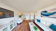 This single-floor residence is designed with a clean, contemporary aesthetic centered around a bright, open-concept living and dining area. Rich wood-look flooring runs throughout the main living space, complementing the crisp white walls and textured ceilings. The living room is generously proportioned, featuring a wall-mounted television, a white storage credenza with decorative glass paneling, and a sliding glass door that fills the room with natural light while providing direct access to an outdoor area.

Adjacent to the living room, the dining area transitions to light tile flooring and accommodates a glass-top table with seating for four. Track lighting overhead provides focused illumination, and a closet offers convenient storage. The kitchen is well-appointed with white shaker-style cabinetry, white countertops, stainless steel appliances including a refrigerator and range, and a double-basin sink beneath a skylight.

The two bedrooms are finished with wood-look flooring and feature louvered closet doors providing ample storage. Natural light enters through standard windows. The bathroom is clean and functional, finished with white tile flooring, a vanity, and a tub-shower combination with a curved curtain rod.