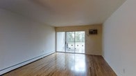 This single-floor space presents a straightforward, functional layout highlighted by gleaming hardwood floors that reflect natural light throughout the main living area. The floors are in excellent condition, adding warmth and a polished aesthetic to the otherwise neutral interior. The walls are finished in a clean, off-white tone, contributing to a bright and open atmosphere.

A large sliding glass door serves as a focal point of the main room, providing direct access to an outdoor balcony enclosed by a metal railing. The door allows generous amounts of natural light to fill the space while offering views of mature trees, creating a pleasant connection to the outdoors.

Climate control is managed by a wall-mounted air conditioning unit positioned adjacent to the sliding door, complemented by baseboard heating units running along the lower perimeter of the walls — a practical setup for year-round comfort. The ceiling is smooth and standard in height, keeping the space feeling tidy and well-proportioned. Overall, the unit presents a clean, move-in ready environment with functional amenities and a calm, neutral palette.