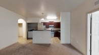 This single-floor space offers a functional and well-appointed open-concept layout that seamlessly connects the kitchen and living areas. The kitchen is a standout feature, showcasing rich dark wood cabinetry that provides ample storage, complemented by stainless steel appliances including a refrigerator, range, and built-in microwave. A central kitchen island adds additional workspace and acts as a natural divider between the kitchen and the adjacent living space. Two pendant lights hang above, casting warm, focused light over the island and enhancing the overall ambiance. The kitchen flooring features a light tile pattern, while the living area transitions to wall-to-wall carpeting, creating a sense of warmth and comfort underfoot. The walls are finished in a clean, neutral tone throughout, contributing to a bright and airy atmosphere. An arched entryway adds an architectural detail that lends character to the space, visible from the main living area. A secondary area or utility space is accessible through a doorway near the kitchen, offering additional practical functionality. Overall, the space combines practical design with comfortable living features.
