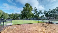 Located at 1660 Old Trolley Rd in Summerville, South Carolina, this single-level outdoor recreational facility offers a well-maintained environment designed for community activity and leisure. The space is anchored by vibrant, color-surfaced tennis courts enclosed within chain-link fencing, providing a dedicated and secure area for play. Towering mature trees, including a prominent hardwood at the center of the grounds, offer natural shade and a lush, park-like ambiance throughout the property. The surrounding landscape features expansive green grass areas that contribute to the open, airy feel of the space. A wood chip ground cover area is present near the courts, suggesting a designated zone for additional recreational use, such as a playground or rest area, complemented by decorative metalwork seating. The combination of well-kept courts, mature canopy trees, and open lawn areas makes this facility a versatile outdoor destination. Set against a backdrop of partly cloudy skies and surrounded by additional tree lines along the perimeter, the space enjoys a peaceful, natural setting while remaining accessible and functional for a variety of outdoor activities.