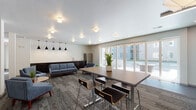 This single-story clubhouse in Memphis, TN (38111) offers a thoughtfully designed communal gathering space with a contemporary aesthetic. The open-plan layout seamlessly connects distinct functional zones, including a relaxed lounge area and a dedicated dining and workspace section. The lounge is furnished with mid-century modern sofas and accent chairs in muted blue-gray tones, arranged around a low round coffee table atop patterned carpet tiles that add textural interest to the floor. A wet bar area with dark cabinetry, a stainless steel refrigerator, and a sink serves as a practical amenity, accented above by a striking row of matte black pendant lights against a textured tile backsplash. The dining and collaborative workspace area features long rectangular tables with modern bar-height seating, illuminated by linear and pendant fixtures. Large multi-panel sliding glass doors along one wall flood the interior with natural light and provide direct access to an outdoor patio area with a pergola structure visible beyond. The flooring transitions between patterned carpet and large-format tile near the entryways, reinforcing distinct activity zones. Neutral white and warm beige walls, recessed and flush-mount ceiling lighting, and carefully placed greenery contribute to the space's clean, welcoming, and functional atmosphere.