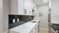 This 2-bedroom, 1-bathroom single-floor residence offers a thoughtfully updated interior with a cohesive modern aesthetic. Light-toned luxury vinyl plank flooring runs throughout the home, creating a warm and uniform foundation across every room. The living room is bright and open, featuring large windows that invite natural light and a sleek ceiling fan overhead. The kitchen is a standout space, boasting crisp white shaker cabinetry with matte black hardware, striking white quartz countertops with bold veining, and a dramatic dark hexagonal tile backsplash. Stainless steel appliances, including a gas range, microwave, and refrigerator, complete the functional and stylish setup. A marble-look peninsula provides additional counter space and defines the transition between the kitchen and living area. The primary bedroom offers a comfortable footprint with a ceiling fan, natural light from a window, and a built-in closet featuring organized shelving and sliding panel doors. The second bedroom is a clean, versatile space with the same warm flooring and neutral walls. The bathroom is finished with marble-style surround tiling in the tub/shower, matte black fixtures and hardware, a wood-toned vanity, and coordinating vinyl flooring, tying together the home's modern design language seamlessly.