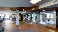 This fitness center is designed to provide a comprehensive workout environment within a single-level layout. The space features an open floor plan with a speckled, rubberized flooring that offers both durability and comfort underfoot. A striking design element is the layered ceiling, which incorporates contrasting tones — a warm rust-brown panel set against a cooler gray surround — adding visual depth to the room. Recessed and flush-mount lighting fixtures, along with a skylight panel overhead, ensure the space is well-lit throughout.

Full-length mirrored walls line one side of the fitness center, visually expanding the space and allowing users to monitor their form during workouts. The equipment selection is varied and includes stationary bikes, elliptical machines, treadmills, and strength-training equipment, catering to both cardio and resistance training needs. A wall-mounted television is positioned for easy viewing during exercise sessions.

Structural columns are integrated throughout the layout, each finished cleanly in white to complement the overall contemporary aesthetic. Natural light filters in through windows visible in the mirrored reflection, contributing to a bright and energizing atmosphere.