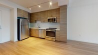 Located in the 90006 corridor of Los Angeles, this single-floor unit presents a contemporary open-concept design that seamlessly connects the kitchen and living areas. Light hardwood-style flooring runs consistently throughout, lending a cohesive, airy feel to the entire space.

The kitchen is well-appointed with a full suite of stainless steel appliances, including a refrigerator, range, built-in microwave, and dishwasher. Textured wood-tone cabinetry extends from floor to ceiling, providing generous storage, while clean white countertops offer ample workspace. Track lighting overhead illuminates the kitchen efficiently. An in-unit washer is conveniently tucked near the entryway.

The open living area benefits from large windows that draw in generous natural light and frame views of the surrounding Los Angeles cityscape. The clean white walls and minimal design create a bright, neutral backdrop adaptable to various furnishing styles.

The bedroom features tall windows fitted with blinds, admitting abundant daylight while maintaining privacy. The bathroom maintains the same modern aesthetic, with a wood-tone vanity, white countertop, large mirror, and light-toned tile flooring that complements the rest of the unit's design palette.