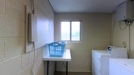 This single-floor laundry facility is built for practicality and efficiency. The room is constructed with painted cinder block walls, giving it a sturdy, utilitarian character typical of a dedicated utility space. A standard-sized window is centrally positioned along one wall, allowing natural light to filter in and offering a view of the surrounding greenery outside, brightening what would otherwise be a purely functional area. A folding table sits at a comfortable working height in the center of the room, providing ample surface area for sorting and folding laundry. To one side, a washer and dryer unit are positioned against the wall, making the most of the available floor space. The smooth, light-colored ceiling contrasts with the textured block walls, contributing to an overall clean and well-maintained appearance. The layout is straightforward and logically organized, ensuring ease of movement and use. This space is designed with functionality at its core, offering everything needed for a complete laundry workflow — from washing and drying to folding — all within a single, well-contained room.