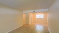 This single-floor space offers a clean and functional layout designed for comfortable everyday living. The main living area is spacious and open, featuring large light-toned ceramic floor tiles that reflect natural light and lend a sense of airiness throughout. Neutral cream-colored walls provide a versatile backdrop, while a textured ceiling adds a subtle layer of character to the interior.

Overhead HVAC vents are integrated into the ceiling, ensuring consistent climate control across the space. A window in the adjoining area allows natural light to filter in, brightening the interior and creating a welcoming atmosphere. The open sightlines connect the living area to what appears to be a kitchen space, which is visible through a wide pass-through opening and includes built-in appliances. A doorway also leads to additional rooms, suggesting a well-organized floor plan that separates living and private areas.

White baseboards and door trim provide clean architectural definition throughout, complementing the overall light and neutral color palette. The layout flows naturally between spaces, making the home feel cohesive and easy to navigate. The combination of tile flooring, ample lighting, and an open plan gives this space a practical yet inviting character suitable for a variety of living arrangements.