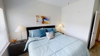 This single-floor space includes a thoughtfully designed bedroom that serves as a restful retreat. The bedroom centers around a generously sized bed dressed in soft blue and teal bedding, accented with decorative pillows in complementary patterns. A dark-toned upholstered headboard anchors the sleeping area, adding a touch of contrast against the clean white walls. Flanking the bed are matching bedside lamps that cast a warm, inviting glow throughout the room, creating a calm and comfortable ambiance. A vibrant abstract canvas featuring bold splashes of orange, blue, and white is displayed above the headboard, providing an eye-catching focal point that adds personality to the otherwise understated palette. The walls are finished in a crisp, bright white that enhances the sense of openness in the room. The overall aesthetic is clean and contemporary, balancing simplicity with carefully chosen decorative elements. The warm lighting, neutral tones, and coordinated furnishings work together to create a serene and welcoming environment well-suited for relaxation.