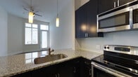 This single-floor space showcases a thoughtfully designed kitchen that serves as a central feature of the home. The kitchen is outfitted with rich, dark cabinetry that extends to the upper walls, providing generous storage while creating a bold visual contrast against the light granite countertops. A mosaic tile backsplash adds texture and a contemporary touch to the cooking area, which is equipped with stainless steel appliances, including a built-in microwave and an electric range. A peninsula with an undermount stainless steel sink and pendant lighting overhead defines the kitchen's open layout, allowing it to connect seamlessly with the adjacent living area. The living space beyond benefits from an abundance of natural light streaming through a set of French doors, which likely open to an outdoor area, enhancing the sense of openness throughout. Elevated ceilings contribute to the airy atmosphere, and a ceiling fan with a light fixture provides both comfort and illumination. The overall design balances modern finishes with a clean, functional aesthetic.