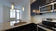 This single-floor space showcases a thoughtfully designed kitchen that serves as a central feature of the home. The kitchen is outfitted with rich, dark cabinetry that extends to the upper walls, providing generous storage while creating a bold visual contrast against the light granite countertops. A mosaic tile backsplash adds texture and a contemporary touch to the cooking area, which is equipped with stainless steel appliances, including a built-in microwave and an electric range. A peninsula with an undermount stainless steel sink and pendant lighting overhead defines the kitchen's open layout, allowing it to connect seamlessly with the adjacent living area. The living space beyond benefits from an abundance of natural light streaming through a set of French doors, which likely open to an outdoor area, enhancing the sense of openness throughout. Elevated ceilings contribute to the airy atmosphere, and a ceiling fan with a light fixture provides both comfort and illumination. The overall design balances modern finishes with a clean, functional aesthetic.