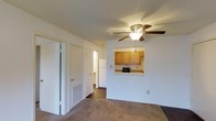 Located at 203 N Cleveland Ave in Long Beach, Mississippi, this single-floor unit offers a practical and comfortable layout designed for easy everyday living. The living area is generously sized, featuring plush wall-to-wall carpeting, a ceiling fan with integrated lighting, and neutral white walls that create a bright, clean atmosphere. A textured ceiling adds a classic touch throughout the space. A standout feature is the pass-through window connecting the living area to the kitchen, which is equipped with warm honey-toned wood cabinetry, a full-size refrigerator, and countertop appliance space. This open connection between the two areas promotes an airy, connected feel. The flooring transitions from carpet in the living space to hard flooring in the kitchen, offering both comfort and practicality. A hallway visible from the living area leads to additional rooms and a bathroom, providing a functional flow throughout the unit. A floor-level air vent ensures efficient climate control. Overall, the space presents a straightforward, well-maintained layout with a cozy ambiance, making it a comfortable and functional home in the Long Beach community.