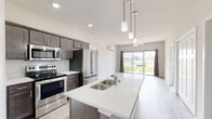 This contemporary single-floor residence showcases a thoughtfully designed open-concept layout that seamlessly connects the kitchen, dining, and living areas. The kitchen is a standout feature, equipped with stainless steel appliances including a refrigerator, range, and over-the-range microwave. Rich dark-toned cabinetry contrasts beautifully with the light quartz countertops and a large central island complete with a double basin sink and stylish pendant lighting overhead. Light wood-look flooring runs throughout the main living areas, enhancing the bright and airy atmosphere. The living area is spacious and comfortable, featuring a ceiling fan, a wall-mounted mini-split unit for climate control, and a sliding glass door that invites natural light and offers outdoor views. The bedrooms are carpeted for warmth and comfort, with generous closet space including a walk-in option. The primary bathroom includes a dual-sink vanity with dark cabinetry and a walk-in shower, while a secondary bathroom offers a single vanity and a soaking tub with safety grab bars. A dedicated pantry area adds practical storage to the kitchen. Throughout, the design maintains a clean, neutral palette that creates a welcoming and modern ambiance.