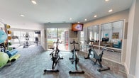 Located at 101 Saint George Blvd in Savannah, Georgia, this well-appointed fitness center offers a comprehensive, single-floor workout facility designed to support a wide range of exercise routines. The space is divided into distinct functional zones, each outfitted with dedicated equipment and thoughtful design elements.

The cardio area features multiple True-brand treadmills, an elliptical trainer, and a recumbent bike, all positioned beneath wall-mounted flat-screen TVs and adjacent to large windows that invite abundant natural light and outdoor views of the surrounding greenery and pool area. A dedicated cycling studio houses a row of stationary spin bikes, also equipped with a mounted TV and direct access to the pool deck through sliding glass doors, creating a bright and energizing atmosphere.

The free weights and functional training zone includes a dumbbell rack and a weight bench, complemented by a well-stocked accessories station featuring medicine balls, stability balls, yoga mats, and resistance bands organized on a convenient rolling rack. Dark cabinetry provides additional storage, and coastal-themed artwork adds a welcoming touch to the décor.

Throughout the facility, patterned teal and gray carpet tiles unify the space while providing cushioning underfoot. Recessed lighting ensures consistent illumination across all zones, contributing to a clean, modern aesthetic suited for an active lifestyle.