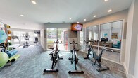 Located at 101 Saint George Blvd in Savannah, Georgia, this well-appointed fitness center offers a comprehensive, single-floor workout facility designed to support a wide range of exercise routines. The space is divided into distinct functional zones, each outfitted with dedicated equipment and thoughtful design elements.

The cardio area features multiple True-brand treadmills, an elliptical trainer, and a recumbent bike, all positioned beneath wall-mounted flat-screen TVs and adjacent to large windows that invite abundant natural light and outdoor views of the surrounding greenery and pool area. A dedicated cycling studio houses a row of stationary spin bikes, also equipped with a mounted TV and direct access to the pool deck through sliding glass doors, creating a bright and energizing atmosphere.

The free weights and functional training zone includes a dumbbell rack and a weight bench, complemented by a well-stocked accessories station featuring medicine balls, stability balls, yoga mats, and resistance bands organized on a convenient rolling rack. Dark cabinetry provides additional storage, and coastal-themed artwork adds a welcoming touch to the décor.

Throughout the facility, patterned teal and gray carpet tiles unify the space while providing cushioning underfoot. Recessed lighting ensures consistent illumination across all zones, contributing to a clean, modern aesthetic suited for an active lifestyle.