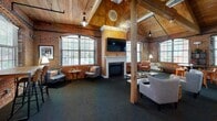Located at 91 Elm St in Manchester, Connecticut, this single-floor space makes a striking architectural statement with its dramatic vaulted wood-plank ceiling supported by heavy timber beams and structural columns. Exposed brick walls run throughout, lending an industrial warmth that pairs seamlessly with the classic white-mantled fireplace, which serves as a natural focal point anchored by a wall-mounted television above. Arched multi-pane windows line the brick walls, flooding the interior with generous natural light while offering views to the surrounding neighborhood. The open living area is thoughtfully arranged with multiple seating groupings — including upholstered armchairs and sofas — centered around a rustic wood coffee table, creating distinct yet connected zones for relaxing and conversation. An orb-style chandelier and vintage-inspired pendant lights complement the industrial aesthetic of exposed ductwork overhead. A kitchenette area with warm wood cabinetry, a sink, and appliances provides practical functionality, while a bistro-style dining table with bar stools adds a casual dining nook. Dark carpet grounds the space and contrasts richly with the warm tones of wood, brick, and natural light throughout.