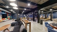 Located at 21 Erie Blvd in Albany, NY, this single-floor entertainment venue offers a generously sized open layout with a raw industrial aesthetic. Exposed concrete ceilings, black-painted structural steel columns, polished concrete floors, and visible ductwork and conduit define the architectural character of the space, lending it a warehouse-style atmosphere that feels both urban and inviting.

The venue is organized into distinct activity zones within the open floor plan. A games area features multiple shuffleboard tables, skee-ball lanes, a billiards table, and a ping-pong table, providing a wide variety of entertainment options. A dedicated lounge zone is anchored by a large sectional sofa, a round coffee table, and colorful accent chairs, all set on a patterned area rug. A bold, multicolored geometric mural serves as a striking focal point on one wall, with a large flat-screen television mounted at its center. A vintage jukebox adds a nostalgic touch nearby.

Additional seating is available at a high-top bar table with wire barstools, positioned near framed blueprint-style artwork. Pendant lighting and recessed track lights illuminate the space throughout. The deep navy blue walls contrast sharply with the raw ceiling overhead, creating a dynamic and energetic ambiance well-suited for social gatherings and events.