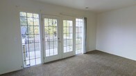 This single-floor space is defined by its bright, open layout and classic architectural details. The main living area is finished with wall-to-wall carpet in a neutral beige tone, creating a warm and comfortable foundation throughout the room. Cream-colored walls complement the light palette, lending the space a clean, airy feel.

A standout feature is the set of white-framed French doors at the center of the room, flanked by matching multi-pane windows that together form a generous wall of glass. This arrangement floods the interior with abundant natural light throughout the day and provides unobstructed views of the surrounding greenery and mature trees. The French doors open directly onto a wooden deck, offering a seamless transition between the indoor living area and the outdoor space.

A curtain rod is installed above the side window, allowing for flexible light control and privacy. The ceiling features a standard height with a visible HVAC vent, ensuring comfortable climate control. Overall, the space presents a simple yet functional design, with its connection to the outdoors serving as a defining and desirable characteristic.