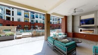 Located at 308 E Hargett St in downtown Raleigh, this single-level covered outdoor lounge serves as a communal amenity space within a modern mixed-use development. The area is defined by a blend of brick columns and smooth painted facades, creating a transitional indoor-outdoor environment that opens directly onto a courtyard and pool deck.

The lounge is organized into distinct seating zones beneath a covered ceiling fitted with ceiling fans, ensuring comfort across seasons. Teal cushioned sofas and accent chairs with metal frames are arranged around low coffee tables, offering a relaxed gathering atmosphere. A built-in linear fireplace is set into a paneled wall alongside a mounted flat-screen television, forming a focal entertainment area ideal for socializing.

Large-format tile flooring extends throughout the covered space and onto the adjacent open patio, which features additional café-style seating and a bar counter with barstools overlooking the pool area. The surrounding multi-story building façade, combining red brick and light panels with large windows, frames the courtyard and contributes to the urban character of the space. Abundant natural light fills the lounge from the open courtyard exposure, enhancing the welcoming and vibrant ambiance of this outdoor retreat.