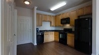This single-floor residence offers a well-designed layout that combines functionality with comfortable living. The kitchen is a standout feature, equipped with warm honey-toned wood cabinetry that extends to the ceiling, complemented by dark granite countertops and a full suite of black appliances, including a refrigerator, range, built-in microwave, and dishwasher. Crown molding adds a refined architectural detail throughout the space, while rich, dark hardwood flooring runs through the kitchen and entry areas, providing a sleek, durable foundation. The kitchen opens directly into the living area, creating a seamless flow ideal for everyday living and entertaining. The living space is finished with plush carpeting and features a ceiling fan with integrated lighting, adding both comfort and practicality. A window with horizontal blinds allows natural light to filter in, casting a warm, inviting ambiance across the room. Crown molding continues into the living area, maintaining a consistent and polished aesthetic. The entry door is a classic paneled design, completing the overall traditional yet modern character of this well-appointed single-floor home.