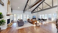 This single-floor community space is defined by its striking architectural character, anchored by soaring vaulted ceilings adorned with bold, dark-stained exposed truss beams that create a dramatic yet welcoming atmosphere throughout. Rich hardwood flooring flows seamlessly across the entire space, complementing the bright white walls and abundant natural light that pours in through large windows and French doors.

The expansive main living area serves as the heart of the space, featuring a statement fireplace with a textured surround, comfortable lounge seating, and a flat-screen television — ideal for relaxed gatherings. Adjacent to the lounge, a dedicated dining area accommodates a large table with modern seating beneath the continued vaulted ceiling, with wide windows offering outdoor views.

A welcoming entryway greets visitors with glass-paneled French doors and a geometric pendant light fixture. An arched doorway leads further into the space, hinting at its thoughtful design details. A front desk area with a dark countertop and retro-style chairs provides a functional service point.

A separate work area features a built-in desk station along the wall, ideal for productivity, alongside a high-top table and direct access to the outdoors via a sliding glass door. Together, these areas create a versatile, well-appointed space that balances communal comfort with practical functionality.