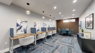 Located at 100 Tuers Ave, this thoughtfully designed co-working space offers a productive and stylish environment for both focused work and collaborative meetings. The single-floor layout is efficiently organized into two functional zones: a row of individual workstations equipped with privacy dividers along one side, and a dedicated group collaboration area anchored by a round table and teal upholstered chairs on the other.

The workstation area features long wooden desks paired with comfortable white chairs, each station separated by fabric privacy panels in a cool blue tone. A series of pendant lights with glass shades hang above, complementing the recessed ceiling lighting to create a bright, well-lit atmosphere. Abstract artwork lines the white walls, adding a creative and professional character to the space.

The collaboration corner is defined by a striking floor-to-ceiling slatted wood accent wall, which introduces warmth and texture against the otherwise clean, neutral palette. A narrow horizontal window above the wood panel allows a touch of natural light to filter into this zone.

Throughout the space, a patterned modular carpet in shades of blue, grey, and beige ties the design together. A wall-mounted split AC unit ensures climate control, while a wooden storage console near the entry provides practical utility. The overall ambiance is modern, calm, and conducive to productivity.
